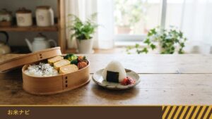 木のテーブルに置かれた、ご飯と卵焼き・ブロッコリー・ミニトマトを詰めたお弁当箱と、おにぎりをのせた小皿
